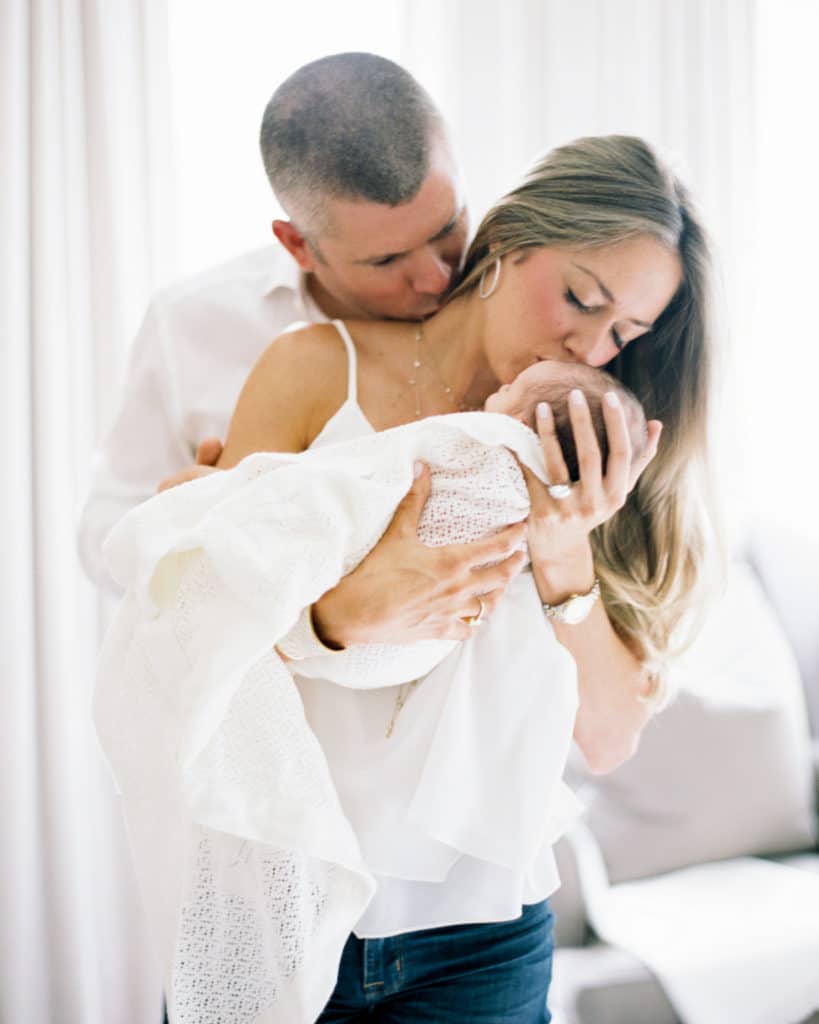 Portrait of mom and dad in an embrace with newborn baby boy in front of window